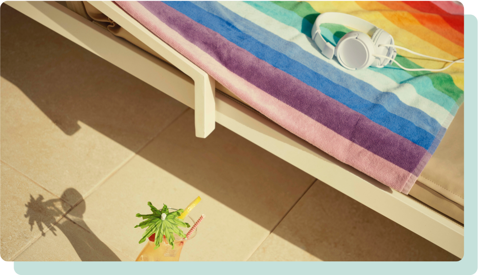 A colorful, striped rainbow towel is draped over a sun lounger, with a pair of white headphones resting on top. Below, on the tiled poolside floor, a tropical cocktail with a green umbrella garnish casts a shadow.
